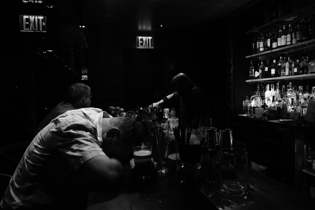Man with head down on bar with alcohol around him in black and white